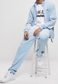 Light blue velour tracksuit with a zip-up jacket and matching pants, featuring a subtle pattern. White sneakers complete the outfit.