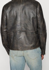 Bruine leren jas met een gestructureerde, versleten uitstraling. Heeft gestikte naden en een iets getailleerde pasvorm, gecombineerd met lichtblauwe jeans.