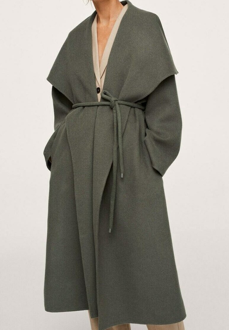 Femme portant un long manteau en laine vert olive ceinturé avec un large col et des poches latérales, sur un blazer et un pantalon beige.