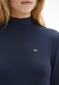 Marinblå ribbad polotröja gjord av ett tyg med mjuk struktur, med en liten broderad logotyp i rött, vitt och blått på bröstet.