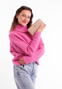 Pink, chunky knit sweater with a high collar, paired with light blue distressed denim. Holding a beige wallet with a dotted pattern and gold logo.