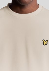 T-shirt beige a girocollo realizzato in tessuto morbido, con un piccolo logo di un'aquila gialla e nera sul lato sinistro del petto.