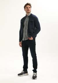 Cardigan navy a maglia con accenti a coste e due tasche anteriori, indossato su una camicia con motivo e abbinato a jeans scuri e sneakers sportive.