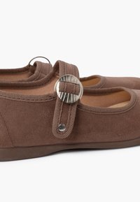 Zapatos de ante marrón con punta redonda, cierre con tira elástica y detalle circular en tono plateado. Forro interior suave visible.