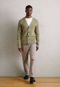 Olivový kardigán s textúrovaným vlnitým vzorom, s predným zapínaním na gombíky, nosený cez biele tričko, v kombinácii so svetlo béžovými nohavicami a čiernymi topánkami.