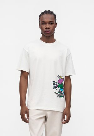Jeune homme aux cheveux tressés portant un T-shirt blanc avec un personnage de dessin animé coloré en train de faire du skateboard sur le côté inférieur.