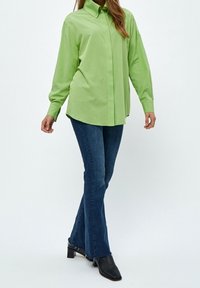 Camicia verde chiaro con bottoni e maniche lunghe, abbinata a jeans svasati blu scuro. Stivali neri con punta a cono completano l'outfit.