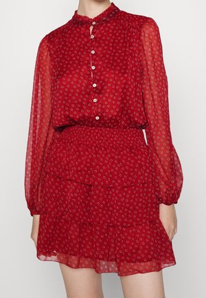 Femme portant une robe rouge à manches longues boutonnée avec de petits imprimés floraux et une jupe à volants à étages sur un fond uni.