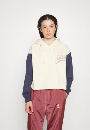 Kuttet hettegenser i krem med marineblå ermer og rosa Adidas-logo. Brukt med rustfargede treningsbukser. Glatt tekstur.