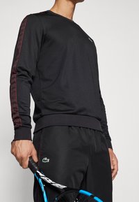 Svart långärmad sweatshirt med röd geometrisk sidopattern, åtsittande ärmslut och en logotyp på bröstet, ihopmatchad med svarta träningsbyxor och ett tennisracket.