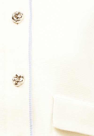 Tissu tricoté crème avec deux boutons décoratifs en forme de nœud argenté à côté d'une couture bleue claire surpiquée et d'une poche à rabat rectangulaire.