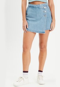 Jupe portefeuille en denim bleu clair avec une texture subtile. Présente une taille haute, des boutons sur le côté et un ourlet asymétrique. Associée à des baskets blanches.