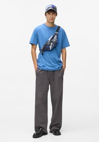 Blå bomulds-t-shirt, løse grå joggingbukser og en mønstret taske. Model bærer en mønstret kasket og sorte slip-on sko. Afslappet outfit.