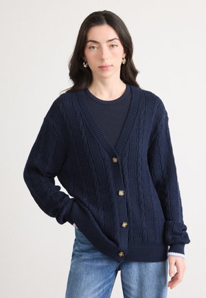 Navy cable-knit cardigan with a V-neck, button closure, and ribbed cuffs, worn over a black top with textured fabric and blue jeans.
