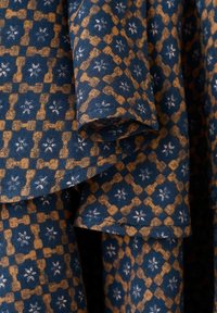 Tessuto blu navy con un motivo geometrico ripetuto di forme marroni collegate e piccoli motivi floreali bianchi, mostrato in morbide pieghe.