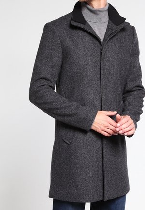 Homme portant un manteau en laine gris foncé par-dessus un col roulé gris clair, les mains jointes devant lui et un pantalon bleu foncé visible.