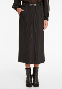 Schwarzer Midirock mit weißen vertikalen Streifen, ausgestattet mit einem Gürtel mit einer metallischen Schnalle, seitlichen Taschen und einem lockeren Schnitt. Getragen mit Stiefeln bis zum Knöchel.