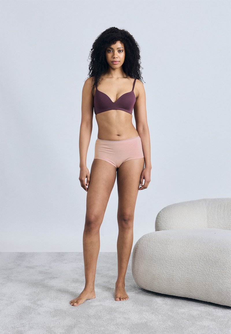 Ασύρματο σουτιέν σε βαθύ μωβ με ομαλή υφή και ντεκολτέ με μεγάλο άνοιγμα, συνδυασμένο με απαλό, ανοιχτό ροζ boyshort. Γυμνά πόδια πάνω σε ανοιχτό χαλί.