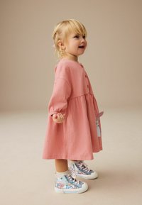 Rosa Langarmkleid mit Knopfverschlüssen und strukturiertem Stoff, kombiniert mit bunten High-Top-Sneakern, die über florale Muster und Spitzenakzente verfügen.