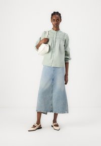 Lys grønn skjorte med smock-detaljer, kombinert med et blått denim maxi-skjørt og beige slip-on sko. Holder en liten hvit veske.