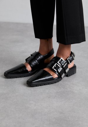 Chaussures slingback en cuir noir avec un bout pointu, comportant des boucles réglables et une semelle épaisse. La finition texturée ajoute du détail.