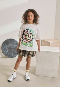 Jeune fille aux cheveux bouclés portant un t-shirt graphique oversized de Blink-182, une jupe à carreaux et des baskets blanches, se tenant à côté d'un cube blanc et d'un accessoire en forme de cercle noir.