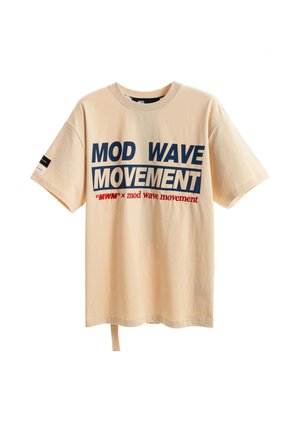 Joven de pie con una camiseta beige "MOD WAVE MOVEMENT", pantalones rojos holgados y zapatos marrones sobre un fondo blanco.