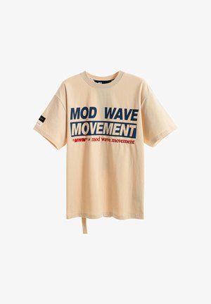 Joven de pie con una camiseta beige "MOD WAVE MOVEMENT", pantalones rojos holgados y zapatos marrones sobre un fondo blanco.