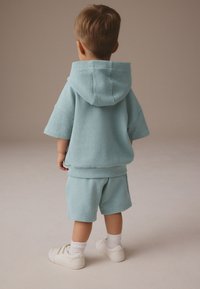 Helehall tekstuuriga lühikeste varrukatega kapuutsi ja samast materjalist lühikeste pükstega komplekt, mille juurde on valitud valged vabaajajalatsid ja pahkluusokid, vaadatuna tagant poolt.