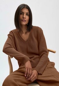 Brauner Strickpullover mit V-Ausschnitt und gerippten Bündchen, kombiniert mit passenden lockeren braunen Hosen, sitzt vor einem neutralen Hintergrund.