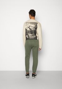 Beiger Pullover mit einem Grafikdruck auf der Rückseite, kombiniert mit olivgrünen Jogginghosen und schwarzen Sneakern. Weicher Stoff; lockere Passform.