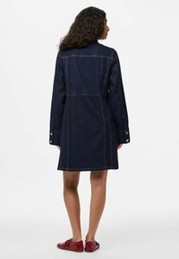 Femme aux cheveux bouclés portant un manteau en denim foncé jusqu'aux genoux et des chaussures plates rouges, se tenant de dos sur un fond blanc.