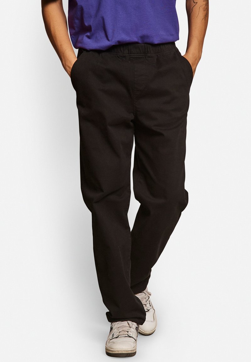 Zwarte, relaxed-fit broek van katoen. Heeft een elastische tailleband, zijzakken en een gladde textuur. G gedragen met een paarse blouse.