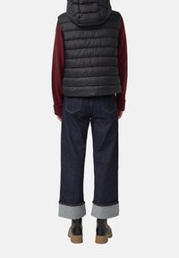 Person in einer schwarzen gepolsterten Weste, burgunderfarbenem Pullover, dunkelblauen, hochgekrempelten Jeans und schwarzen Stiefeln, der mit dem Rücken zur Kamera vor einem weißen Hintergrund steht.