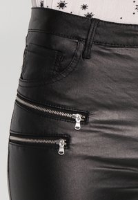 Pantaloni neri in faux pelle con una texture liscia, cinque tasche e due dettagli con zip frontale. Le zip in metallo hanno hardware argentato.