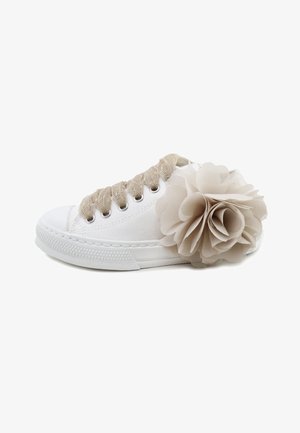 Zapatilla de lona blanca con cordones beige y una gran flor de tela beige pegada cerca del talón sobre un fondo blanco.