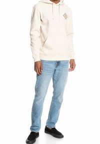 Svetle béžová mikina s kapucí, šňůrkami a přední kapsou, se diamantovým logem na prsou; doplněno modrými džíny a černými botami.