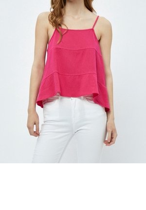 Vrouw die een mouwloze, loszittende roze top met dunne bandjes draagt, gecombineerd met een witte broek met hoge taille, tegen een effen achtergrond.