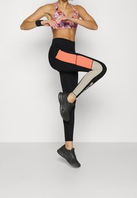 Leggings sportivi neri con pannelli arancioni e beige. Il modello li abbina a un reggiseno sportivo floreale multicolore e a scarpe da corsa nere.