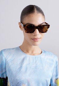 Lunettes de soleil en écaille avec un design géométrique et des verres foncés. Le modèle porte une chemise bleu clair, texturée, avec des motifs subtils.