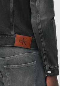 Čierna džínsová bunda so svetlosivým prešívaním, s hnedou koženou štítkom s vyrazeným logom a striebornými gombíkmi na rukávoch.