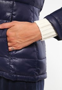 Granatowa kurtka puchowa z zamykaną na zamek kieszenią, błyszczącą fakturą i białymi ściągaczami z dzianiny, podkreślająca ciepło i funkcjonalność.