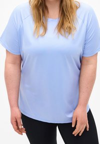 Mujer vestida con una camiseta de manga corta azul claro y pantalones negros, de pie con las manos relajadas a los lados, recortada a la altura de los hombros y los muslos.