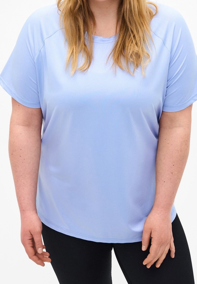 Mujer vestida con una camiseta de manga corta azul claro y pantalones negros, de pie con las manos relajadas a los lados, recortada a la altura de los hombros y los muslos.