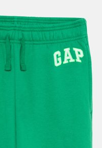Zaļas sporta bikses ar elastīgu jostu, auklu un izšūtu "GAP" logotipu gaišākā zaļā krāsā. Gluda auduma faktūra.