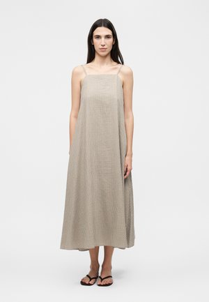 Woman standing wearing a beige and white striped sleeveless midi dress with thin straps and black sandals on a white background.