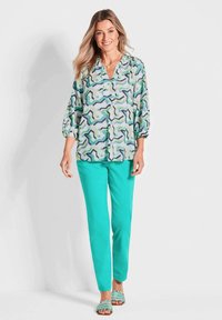 Leichte, gemusterte Bluse mit blauen, grünen und schwarzen abstrakten Formen, kombiniert mit einfarbigen, türkisfarbenen Slim Pants und mit Perlen verzierten Sandalen.