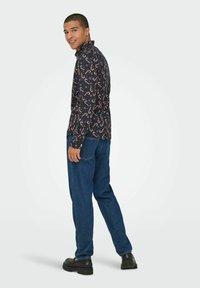 Noor mees naeratab, kandes musta pika varrukaga särki, millel on piparkoogikarbi muster, siniseid teksaseid ja musti kingi, seistes lihtsa heleda tausta ees.