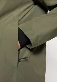 Jachetă impermeabilă de culoare verde măslin cu un design elegant, având o buzunar ascuns, manșete elastice și detaliu cu șnur la tiv.