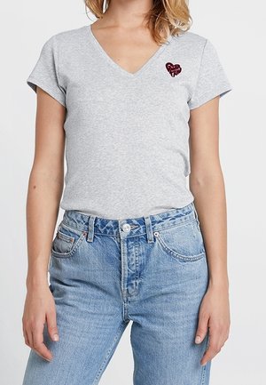 Femme portant un T-shirt gris clair à encolure en V avec un petit cœur rouge et un jean taille haute bleu clair, debout devant un fond uni.